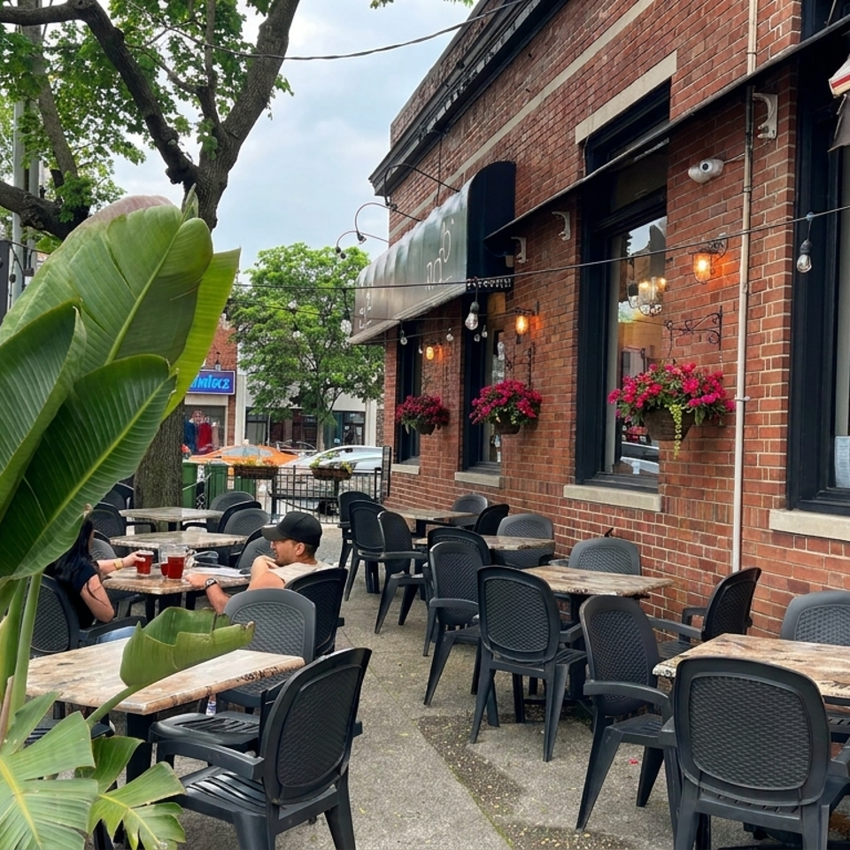 Outdoor restaurant patio with brick walls, black chairs, wooden tables, hanging flowers, string lights, and tropical plants.